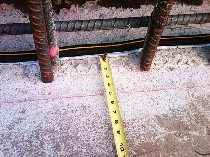 Close-up of a measuring tape on a concrete surface, with exposed rebar and a black rubber strip nearby, including a faint red line marking on the concrete.