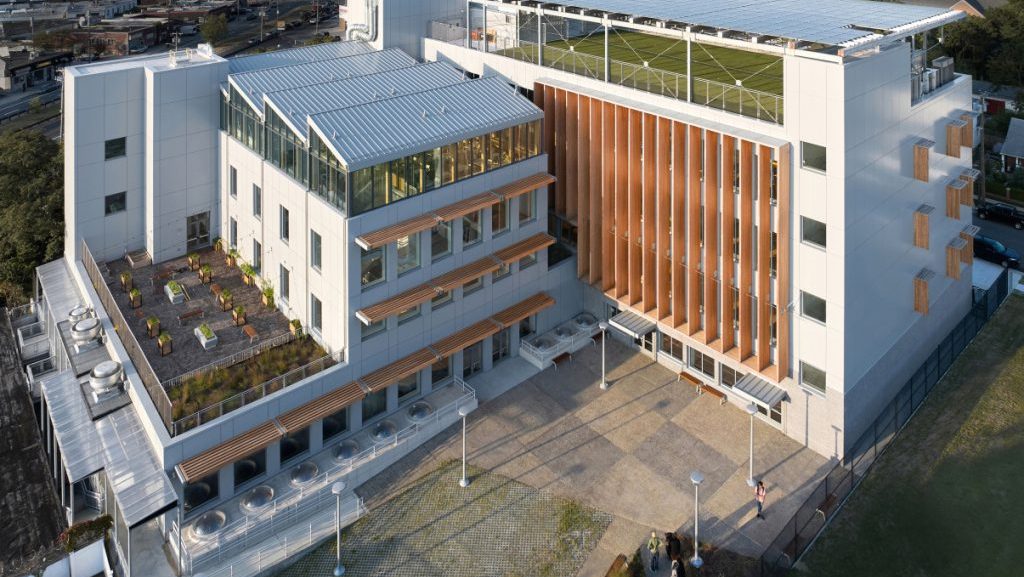 Aerial view of a modern white institutional building with rooftop artificial-turf fields, landscaped terraces, and a paved courtyard in an urban setting.