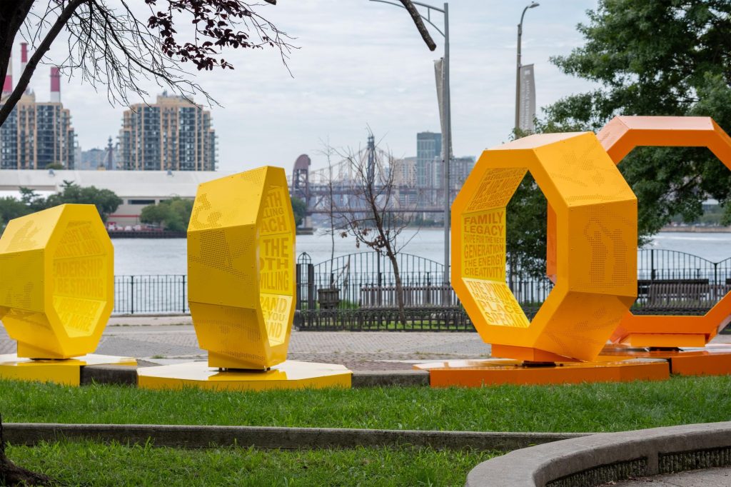 Bright yellow-orange geometric sculptures with cutout lettering stand beside a river, with trees, a bridge, and high-rise buildings in the background.