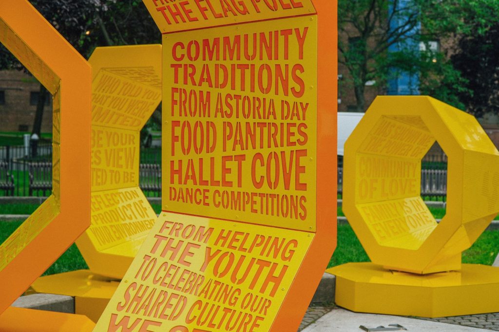 Yellow-and-orange geometric outdoor sculpture with red text about community traditions and activities in a tree-lined park.