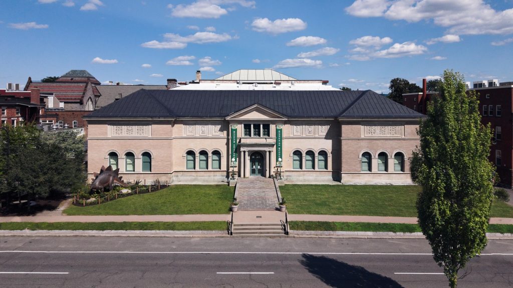 Front view of the Berkshire Museum, a historic brick building with a dark roof, green banners, and a dinosaur sculpture on the lawn.
