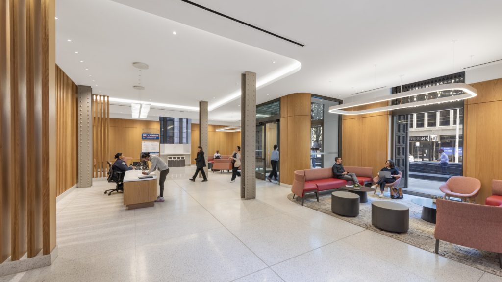 Modern wood-paneled lobby with a reception desk, people, lounge seating, and large street-facing windows.