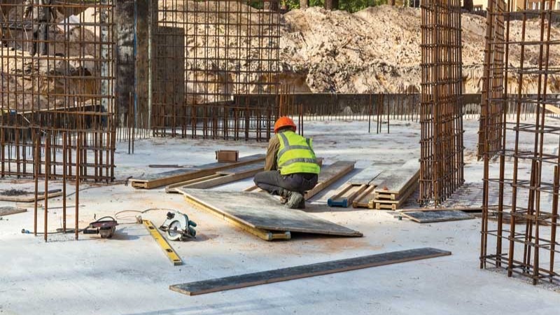 Construction worker in a high-visibility vest kneeling on a concrete slab amid rebar columns and scattered building materials.
