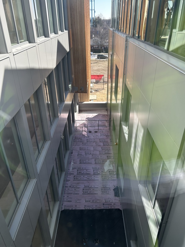 View down a narrow construction space between two modern buildings, with insulation-covered flooring, reflective windows, and a residential street visible beyond.