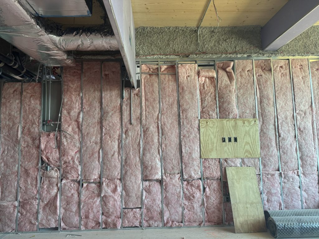Unfinished interior wall with pink insulation, exposed electrical wiring, HVAC ductwork, and a plywood panel containing three electrical boxes.
