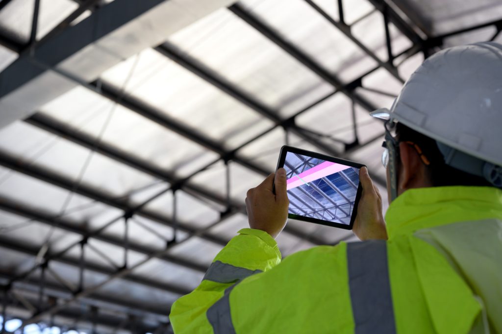 Worker in a hard hat uses a tablet to inspect a glass-and-metal roof structure.