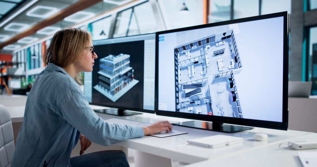 Woman reviewing a 3D building model and architectural floor plan on two computer monitors.