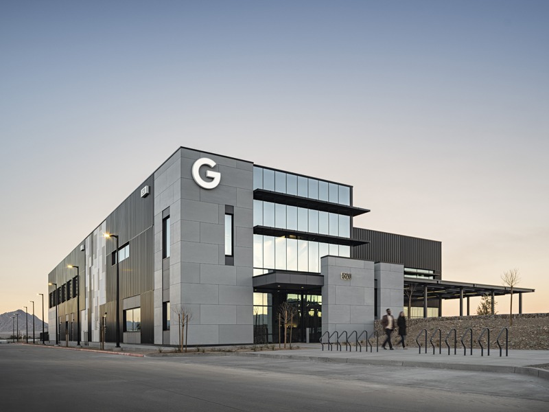 Modern gray-and-glass building with a large “G” sign, bicycle racks, pedestrians, and mountains in the background.