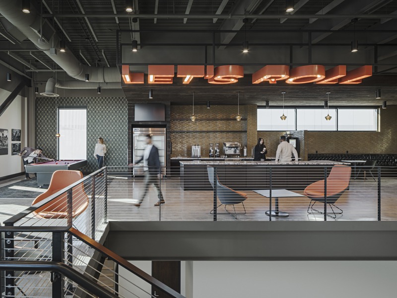 Industrial-style café or lounge with a bar, pool table, colorful chairs, exposed ceiling, and staircase railing.