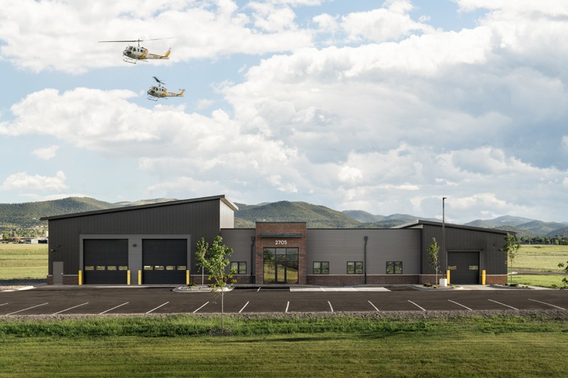 Dark gray industrial building with a parking lot, two helicopters overhead, and green mountains in the background.