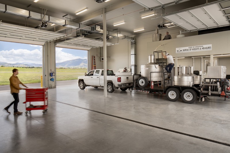 Industrial Bureau of Weights & Measures facility with a pickup truck, equipment trailer, worker, and person pushing a red tool chest; mountains are visible through the open garage doors.