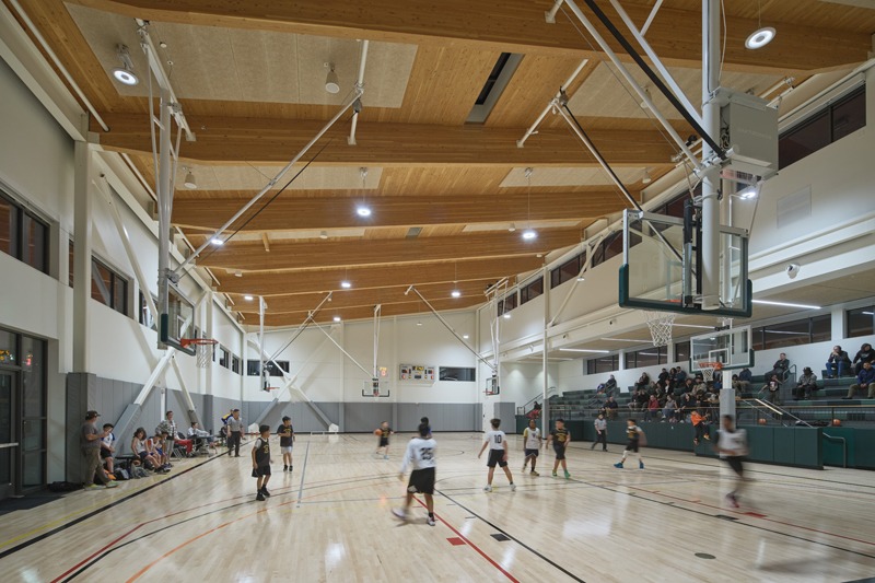 Players compete in a basketball game inside a brightly lit gymnasium with spectators seated on green bleachers.