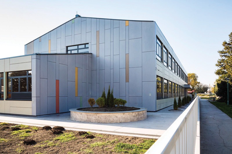 Modern gray-paneled building with colorful vertical accents, large windows, a curved planter, and a walkway bordered by a white railing.