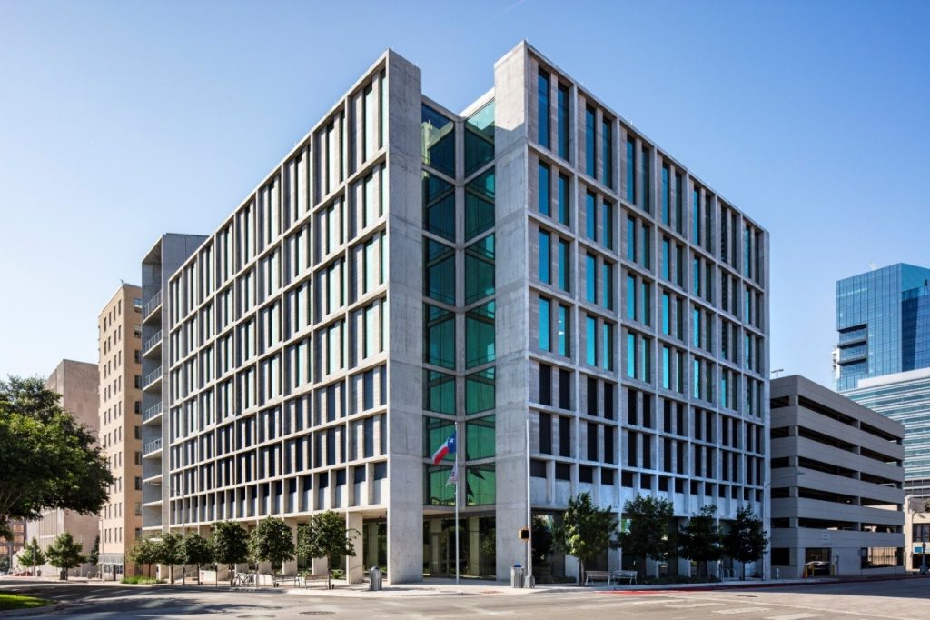Modern concrete-and-glass office building with a central glass atrium, flags, street trees, and surrounding downtown buildings.