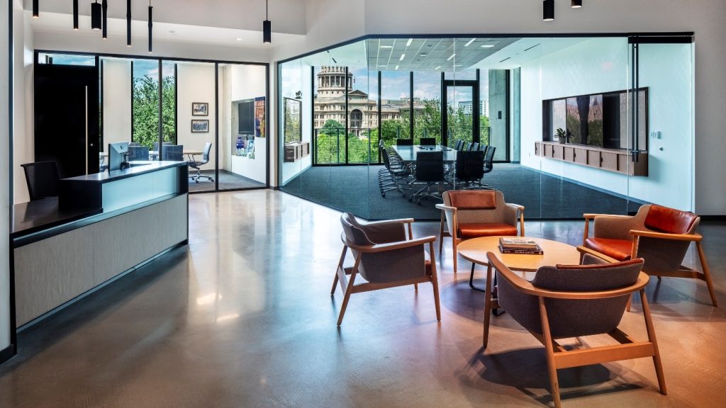 Modern office lobby with a reception desk, glass-walled conference room, lounge seating, and a view of a historic tower through the windows.