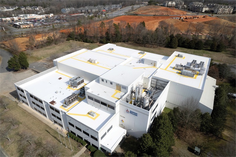 Aerial view of a large white industrial building with rooftop mechanical equipment and yellow markings, bordered by trees and a red-earth construction site.