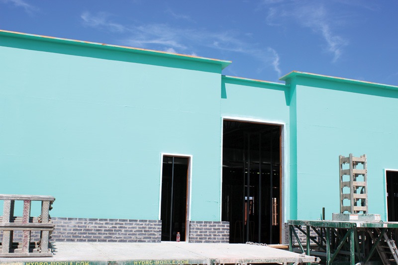 Building under construction with walls coated in bright teal air barrier. Several dark door and window openings are framed in white, with gray brick starting at the base. Scaffolding stands in the foreground against a blue sky.