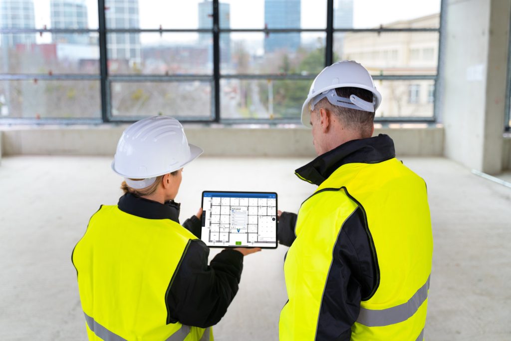 Two construction workers in hard hats and high-visibility vests review a digital floor plan on a tablet inside an unfinished building.