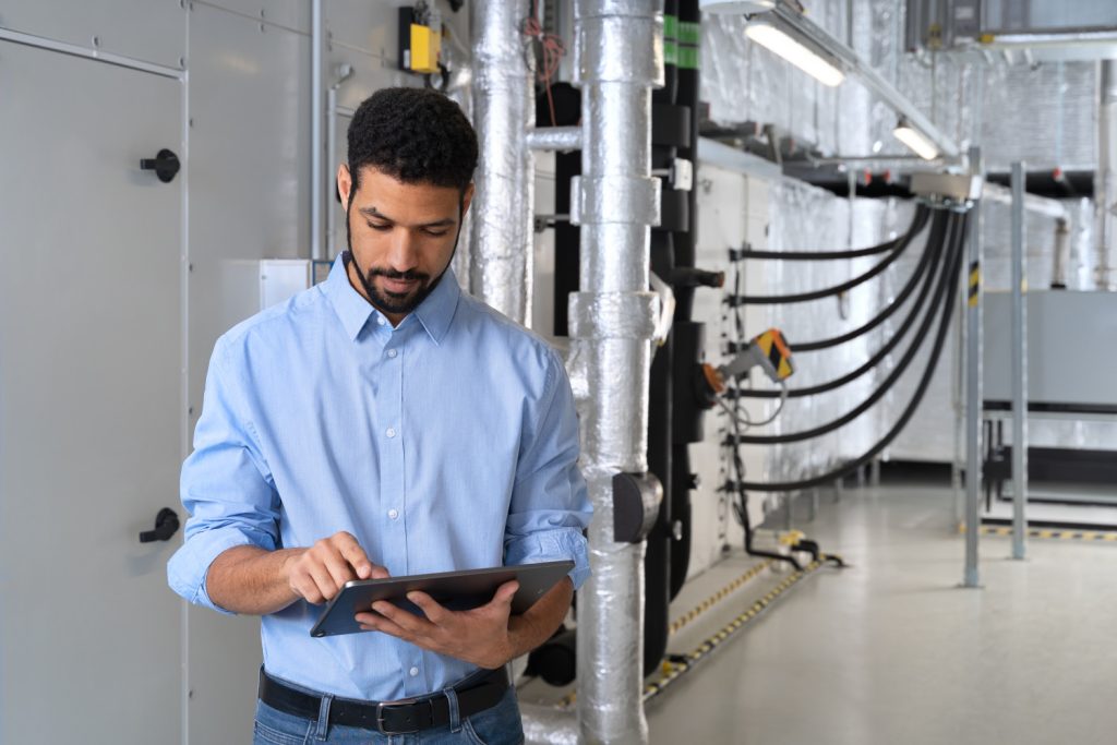 Man using a tablet in an industrial mechanical room with insulated ducts and equipment.