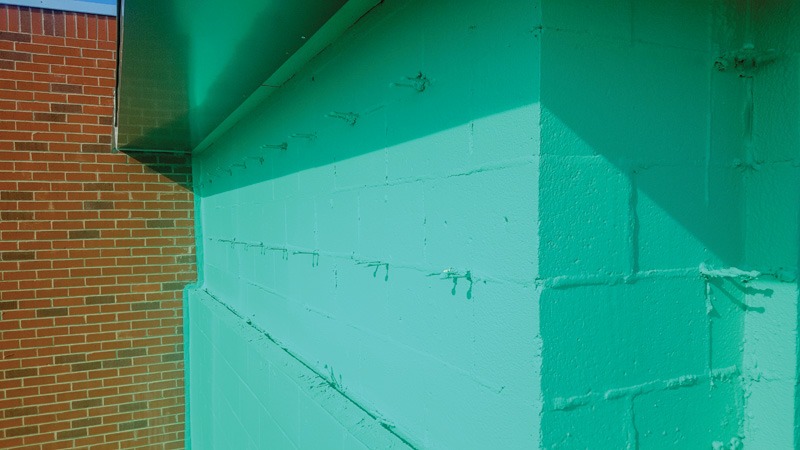 Corner of a concrete block wall coated in teal-green fluid-applied air barrier, with a red brick wall and a metal flashing overhang at the upper left. Drips and ridges from the coating show along the mortar joints.