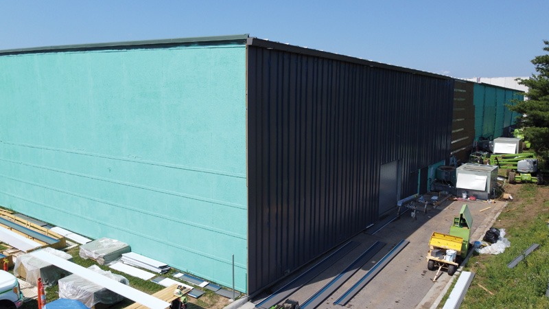 Exterior of a large industrial building under construction. The left half of the wall is coated in teal air barrier, and the right half has dark charcoal vertical metal panels already installed. Construction equipment, stacked materials, and trim pieces sit on the ground in front.