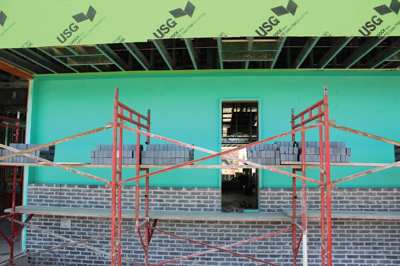 Teal air-barrier-coated wall with a narrow window opening above a partially built gray brick base. Red scaffolding holds stacks of brick, and green USG sheathing panels hang overhead.