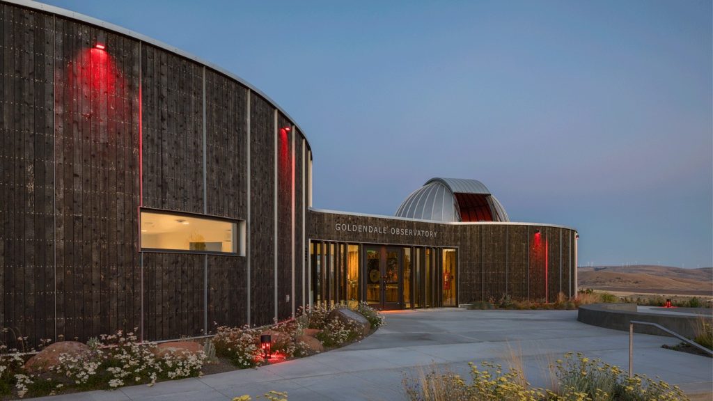 Modern Goldendale Observatory building with dark curved wooden walls, red lights, a domed telescope enclosure, and a paved landscaped entrance at dusk.