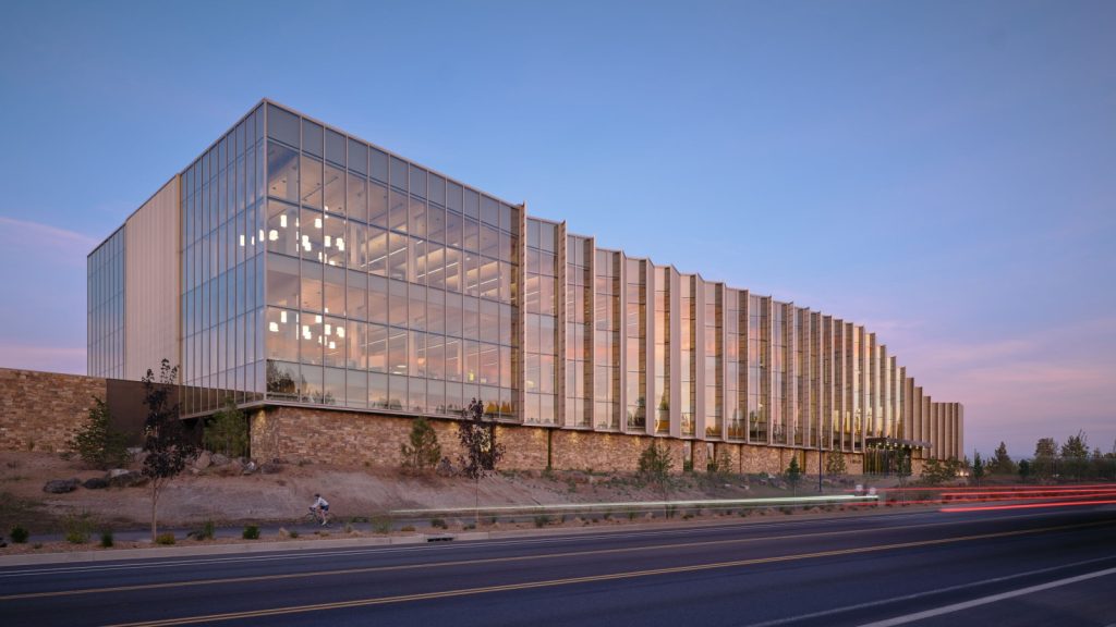 Modern glass-fronted library illuminated at dusk beside a multilane road.