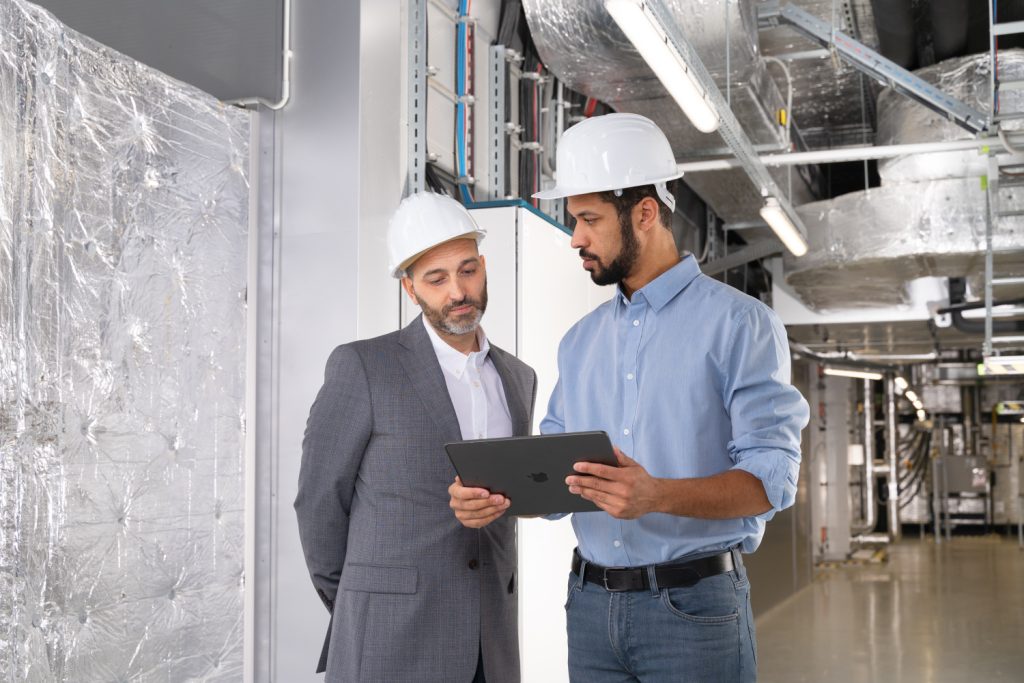 Two men in hard hats examine a tablet inside an industrial facility.