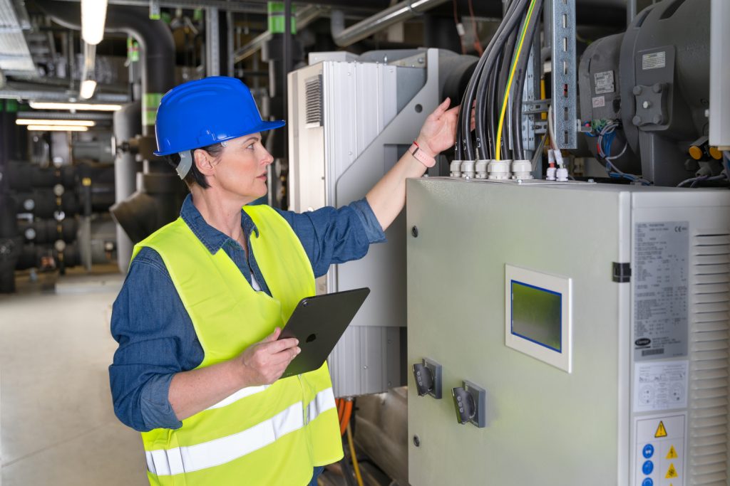 Worker in a hard hat and reflective vest inspects an electrical control cabinet while holding a tablet.