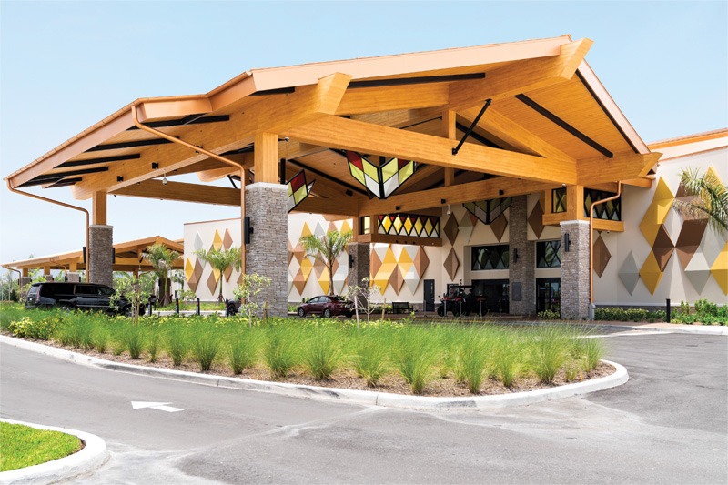 Modern building entrance with a large wooden porte-cochère, geometric multicolored wall decorations, parked cars, and landscaped driveway.
