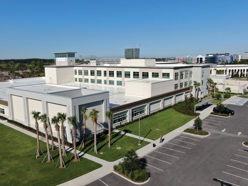 Aerial view of a modern white building complex with palm trees, landscaped grounds, and a parking lot.