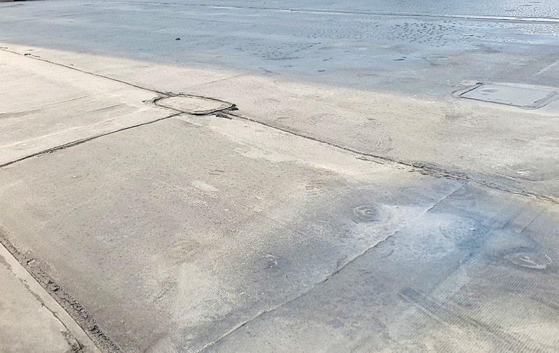 A weathered concrete surface with seams, utility covers, cracks, and shallow reflective puddles.