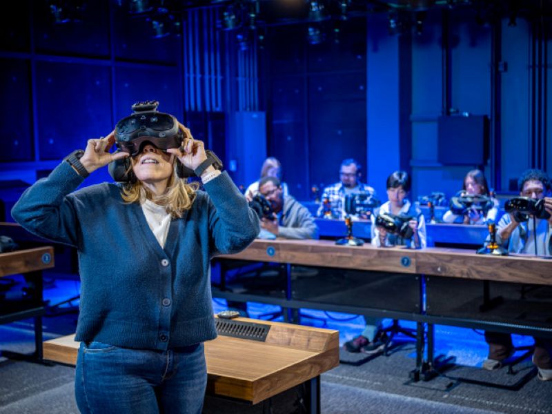 Woman wearing a virtual-reality headset in a blue-lit technology lab while people work at tables behind her.